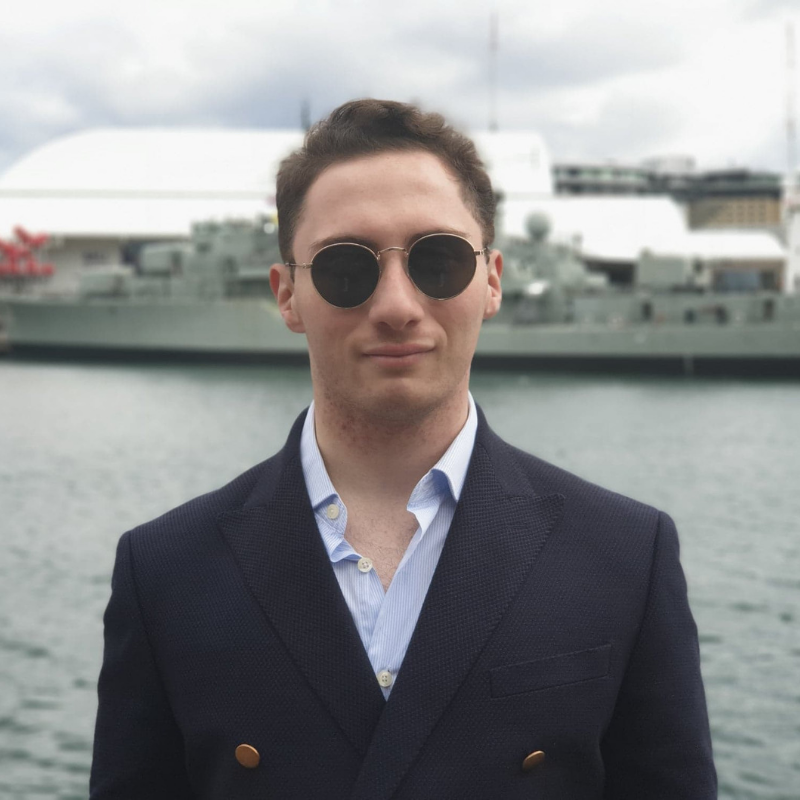 Photo of Ryan Attard. He is in a suit and glasses in front of a boat.